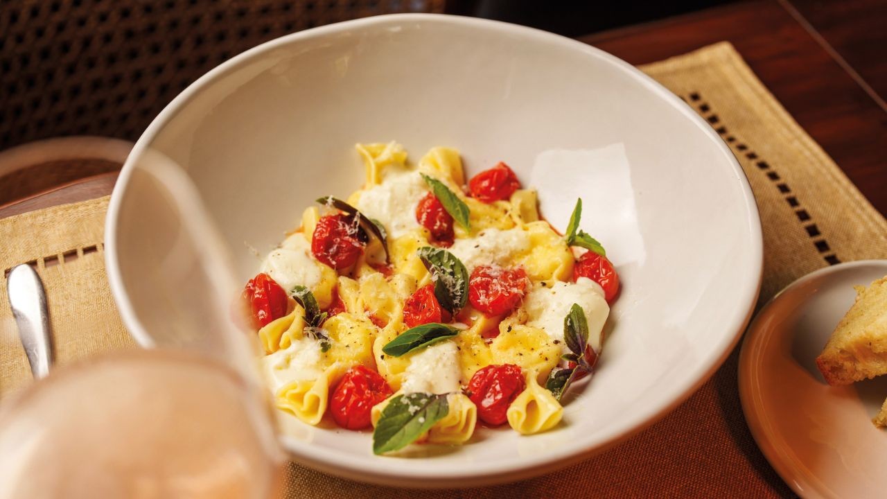 Massa tortellini amarela com molho branco, tomates cereja vermelhos e folhas de manjericão verde em uma tigela branca, sobre um jogo americano marrom