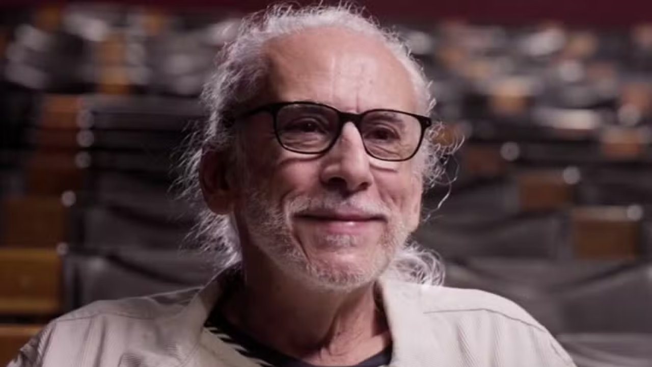 Tulé Peake: homem idoso de pele clara, cabelos e barba grisalhos, usando óculos de armação preta e camisa clara, sorrindo levemente para a câmera
