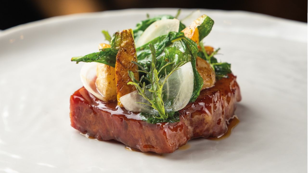 Prato gourmet com um pedaço de carne bovina caramelizada, coberto por folhas verdes, fatias de cebola roxa, batatas rústicas e um raminho de alecrim, servido em um prato branco