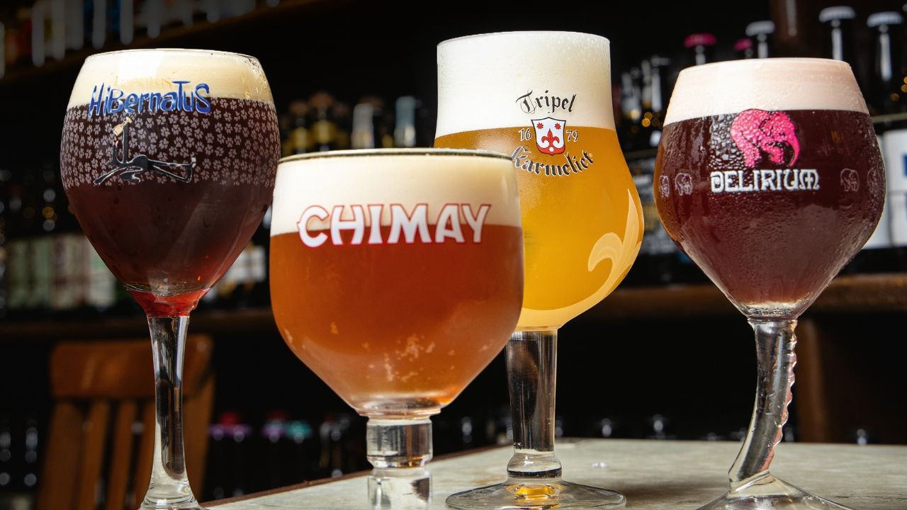 Quatro copos de cerveja com diferentes cores e espumas, da esquerda para a direita: uma cerveja escura com "Hibernatus", uma âmbar com "Chimay", uma dourada com "Tripel Karmeliet" e uma vermelha com "Delirium" e um elefante rosa, sobre um balcão com garrafas desfocadas ao fundo