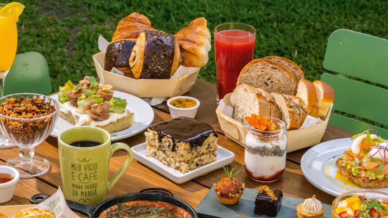 Mesa de madeira com café da manhã variado ao ar livre, incluindo pães, croissants, bolo de chocolate, sucos, iogurte com granola e torradas com abacate e salmão, com gramado verde ao fundo