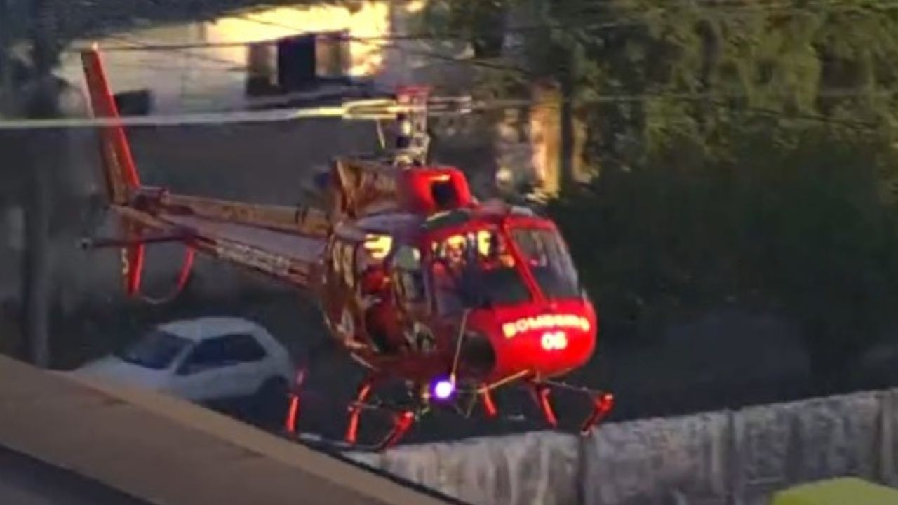 Helic&oacute;ptero vermelho dos Bombeiros, com a inscri&ccedil;&atilde;o BOMBEIROS 06 na frente, pairando baixo sobre uma &aacute;rea urbana com casas e &aacute;rvores ao fundo, e um carro branco estacionado abaixo. A luz roxa da aeronave est&aacute; acesa, indicando opera&ccedil;&atilde;o noturna ou de baixa visibilidade