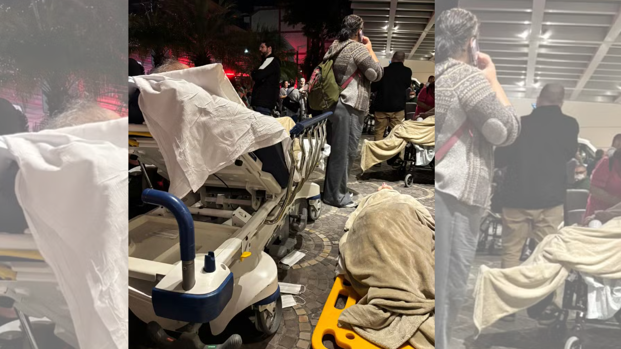 Pessoas em um ambiente noturno, possivelmente um hospital ou abrigo, com macas e cobertores. Uma mulher de casaco cinza e mochila marrom está de costas, enquanto outras pessoas e equipamentos médicos são visíveis ao fundo. Máscaras descartáveis estão no chão, indicando um cenário de emergência ou aglomeração.