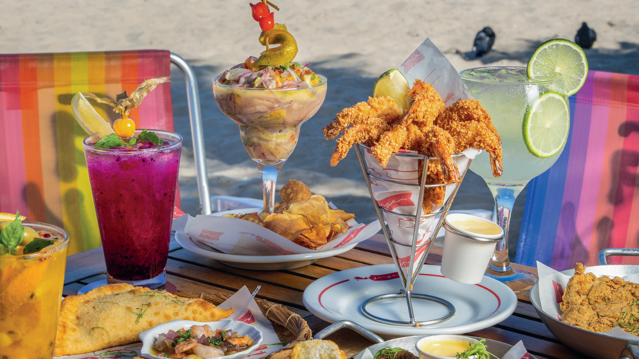 Mesa de madeira com diversos pratos e bebidas coloridas na praia. Há um drink roxo com physalis, um ceviche em taça, camarões fritos, pastéis, e um drink verde com limão. Cadeiras de praia listradas e areia ao fundo