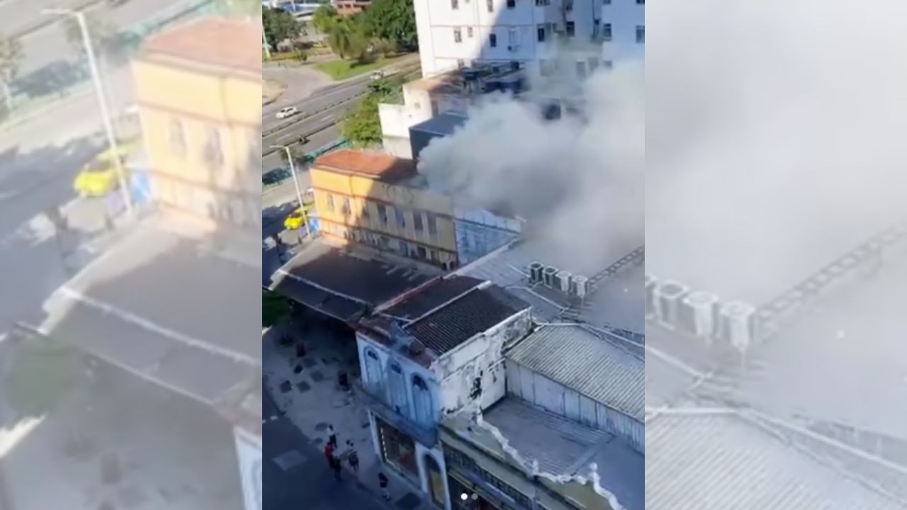Vista aérea de um prédio amarelo de dois andares com telhado marrom, liberando fumaça cinza escura de suas janelas e telhado. Abaixo, outros prédios com telhados escuros e pessoas na calçada. Ao fundo, uma rua movimentada com carros e árvores. O céu está claro e ensolarado