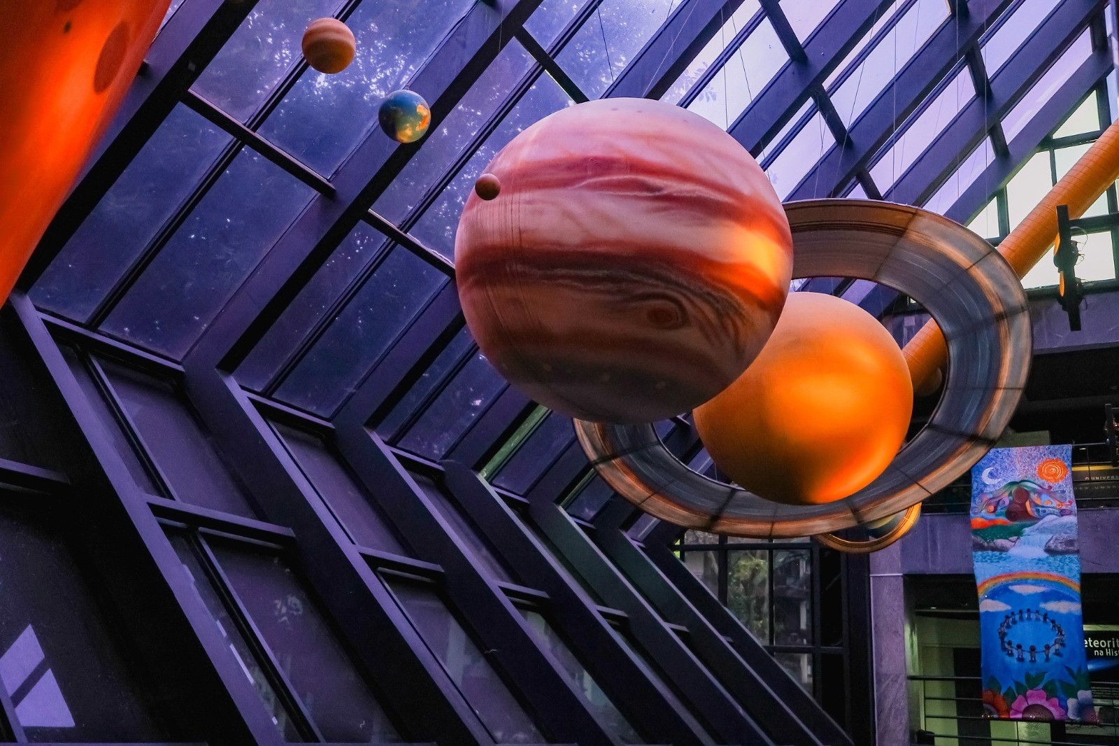 Modelos de planetas suspensos em um ambiente interno com teto de vidro. J&uacute;piter, com suas faixas marrons e laranjas, est&aacute; em primeiro plano, ao lado de um planeta laranja sem an&eacute;is e outro com an&eacute;is prateados. Mais ao fundo, pequenos modelos da Terra e de Marte s&atilde;o vis&iacute;veis. O teto inclinado &eacute; composto por pain&eacute;is de vidro e estruturas met&aacute;licas escuras. No canto inferior direito, uma bandeira colorida com desenhos de paisagens e pessoas. A ilumina&ccedil;&atilde;o &eacute; difusa, com tons roxos e alaranjados
