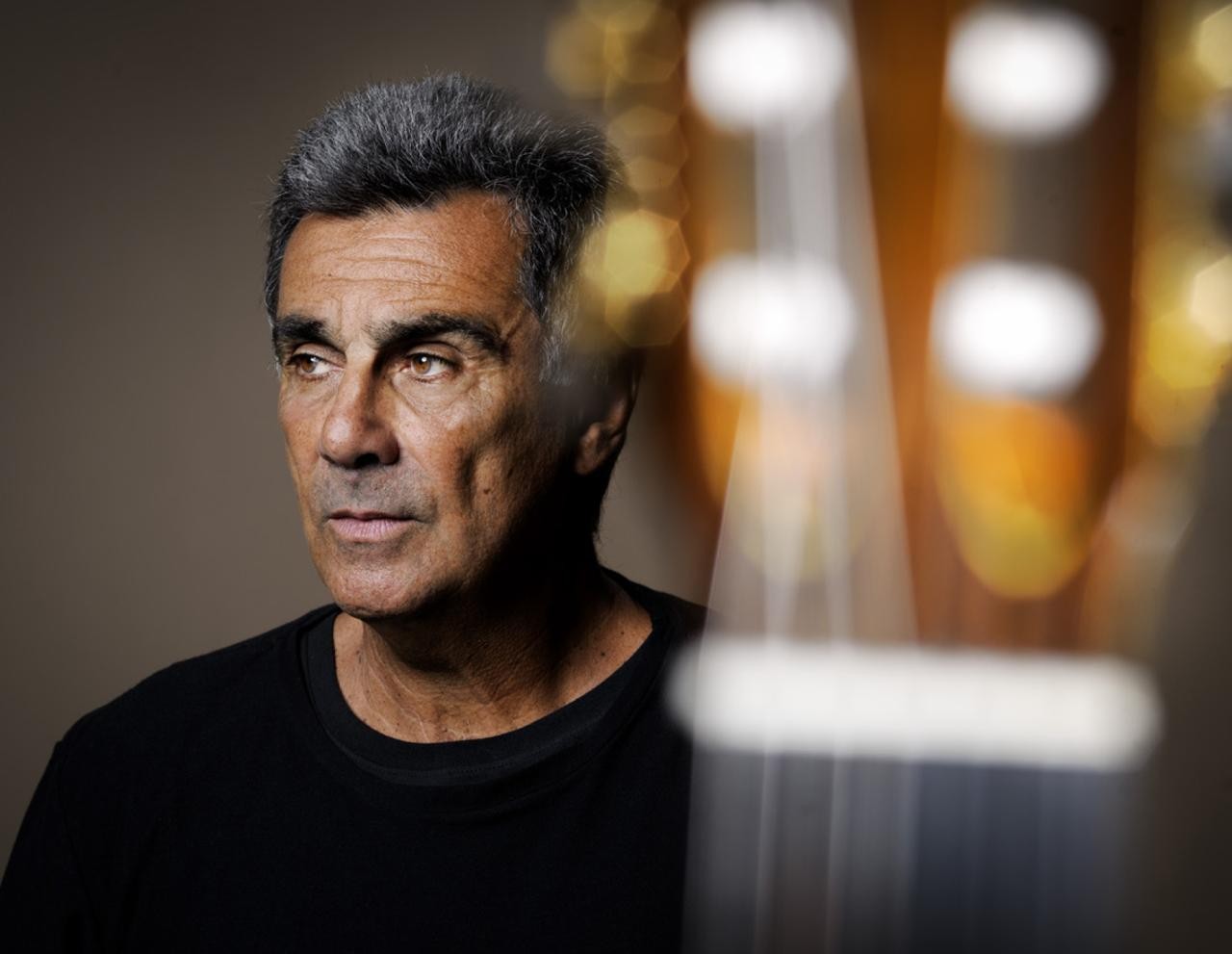 Homem de pele bronzeada, cabelo grisalho e camiseta preta, olhando pensativo para a esquerda, com um instrumento musical desfocado &agrave; direita