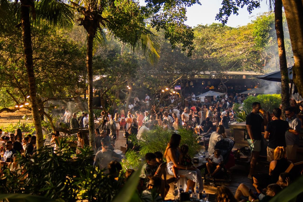 Multid&atilde;o de pessoas em um evento ao ar livre, com palmeiras e vegeta&ccedil;&atilde;o densa. H&aacute; fuma&ccedil;a no ar, luzes penduradas e barracas, sugerindo uma festa ou festival em meio &agrave; natureza