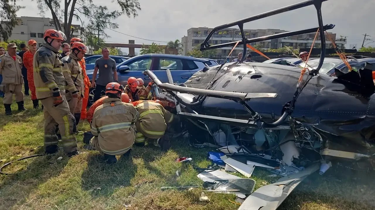 Bombeiros em uniformes de resgate laranja e bege inspecionam um helicóptero preto acidentado, com a fuselagem danificada e partes espalhadas na grama. Ao fundo, carros e edifícios sob céu claro