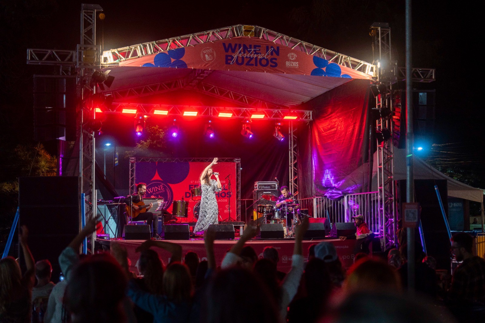 Cantora de vestido estampado no palco iluminado por luzes vermelhas e roxas, com banda ao fundo, durante o evento 