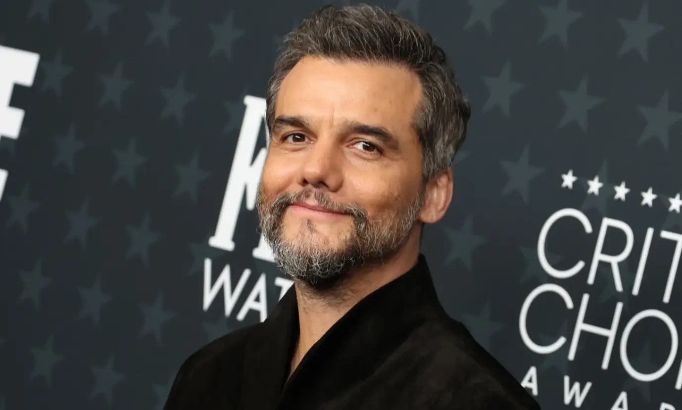 Wagner Moura sorrindo levemente, com barba e cabelo grisalhos, vestindo uma camisa preta, em frente a um fundo escuro com estrelas e o texto CRITICS CHOICE AWARDS