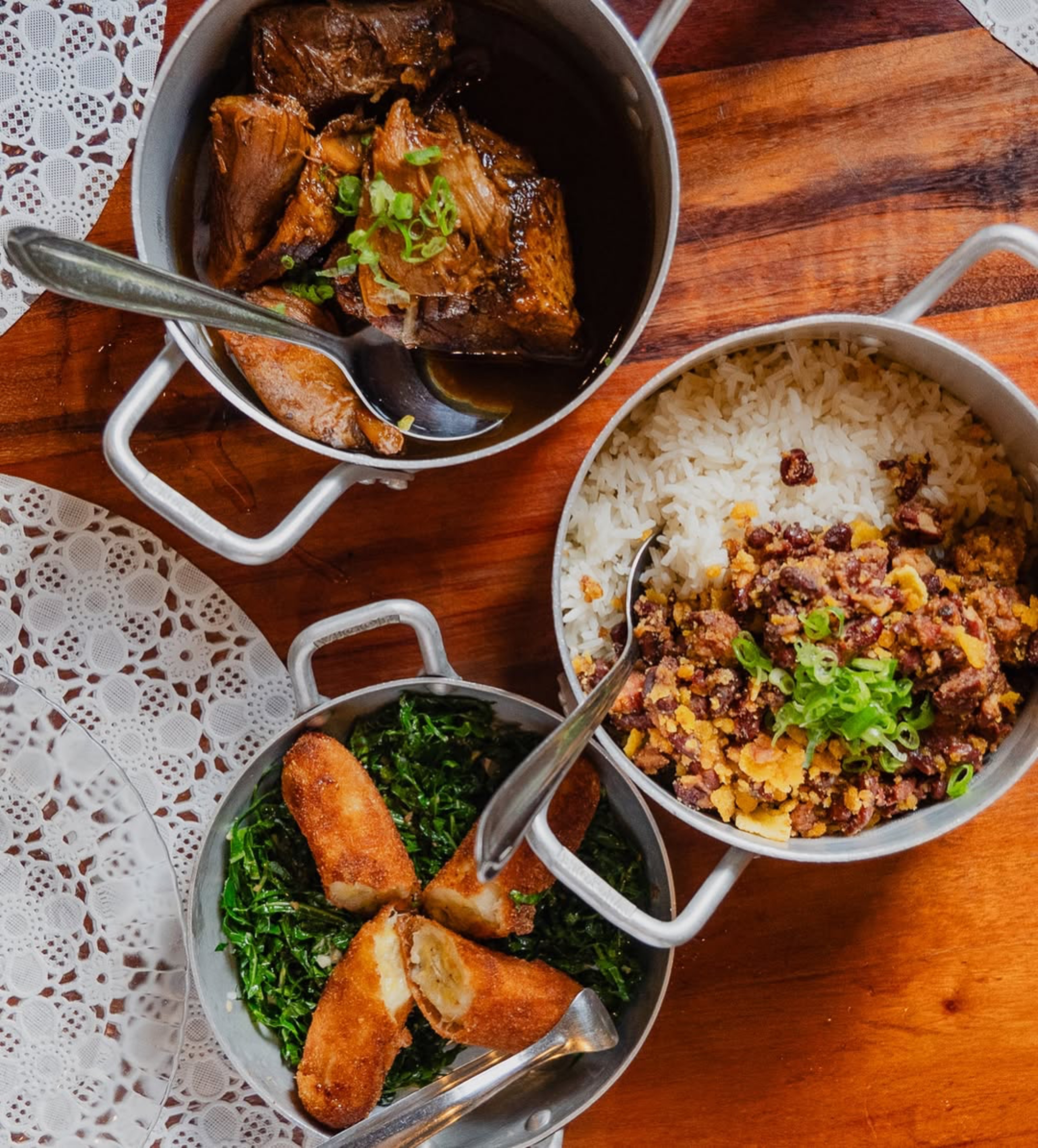 Tr&ecirc;s panelas de alum&iacute;nio com comida brasileira sobre uma mesa de madeira. A panela superior tem carne de panela com cebolinha, a do meio tem arroz branco com feij&atilde;o tropeiro e a inferior tem couve refogada com banana &agrave; milanesa
