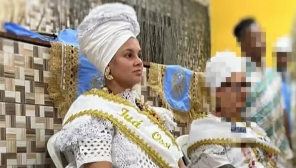 Mulher de turbante branco e roupa de renda branca com faixa dourada Iya Osi no peito, sentada, olhando para a direita, com uma criança ao lado e outras pessoas desfocadas ao fundo