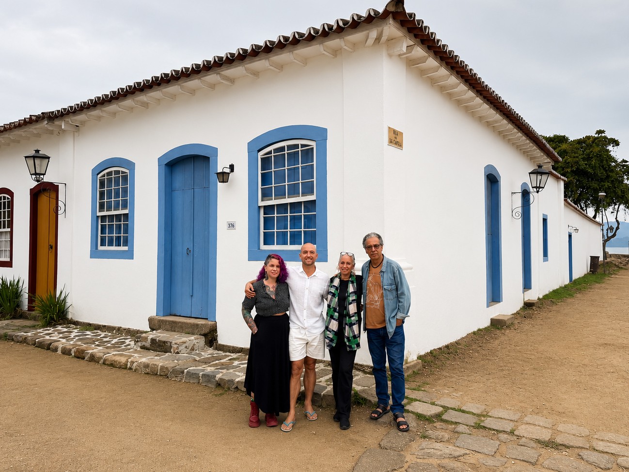 Quatro pessoas sorridentes posam em frente a uma casa colonial branca com portas e janelas azuis em Paraty. Da esquerda para a direita: uma mulher de cabelo roxo e saia preta, um homem de camisa branca e bermuda, uma mulher de cabelo grisalho e cachecol xadrez, e um homem de &oacute;culos e jaqueta jeans. O ch&atilde;o &eacute; de terra e pedras, com o mar vis&iacute;vel ao fundo