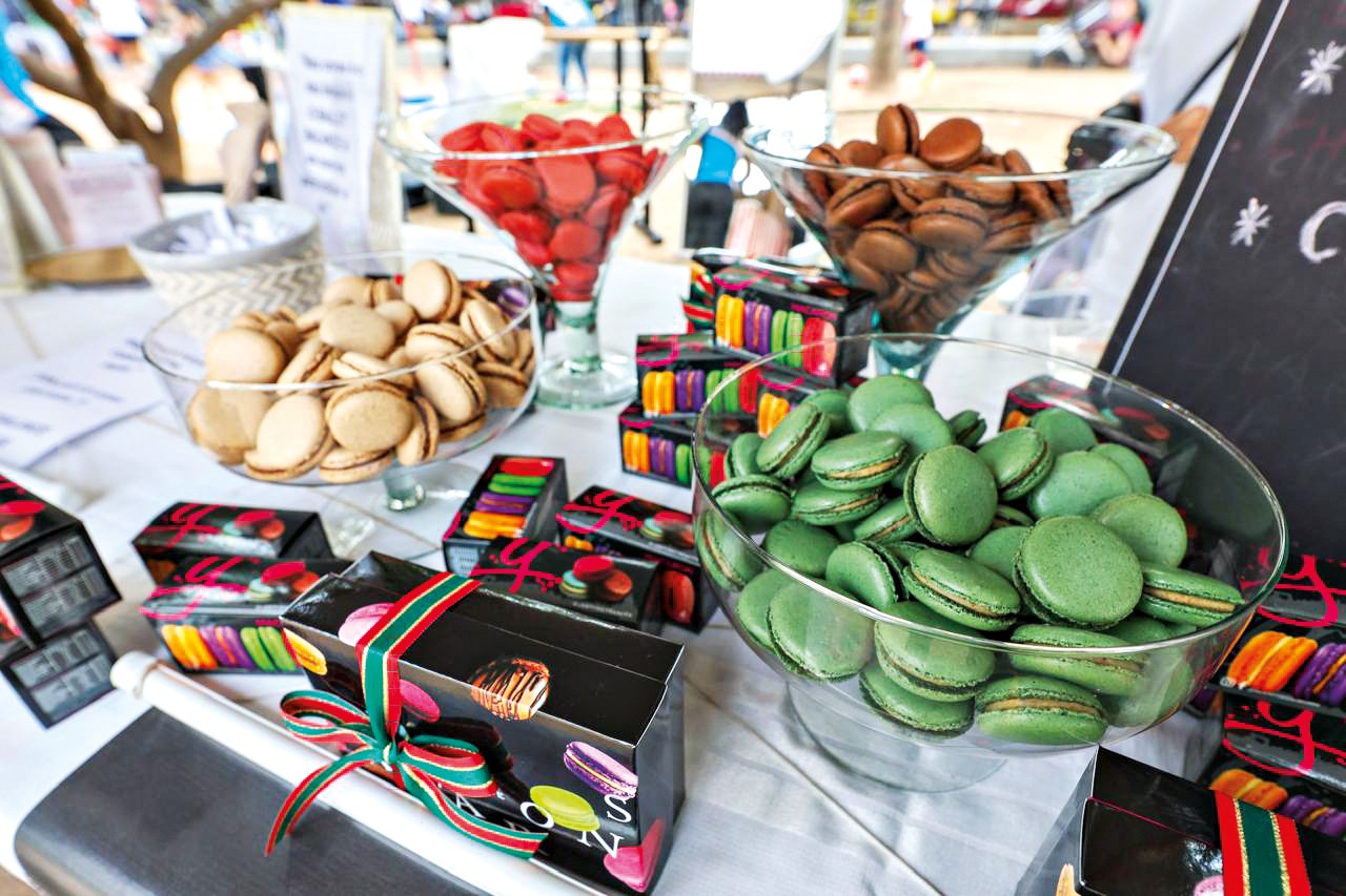 Mesa com tigelas de vidro cheias de macarons coloridos: bege, vermelho, marrom e verde. H&aacute; caixas pretas com la&ccedil;os coloridos e estampas de macarons, e outras caixas com fileiras de macarons coloridos. Um quadro-negro com escrita branca aparece &agrave; direita