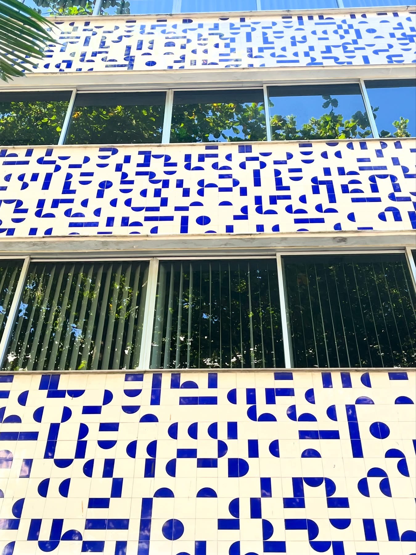 Fachada de edifício com azulejos brancos e padrões geométricos azuis, intercalados por janelas escuras que refletem árvores e o céu