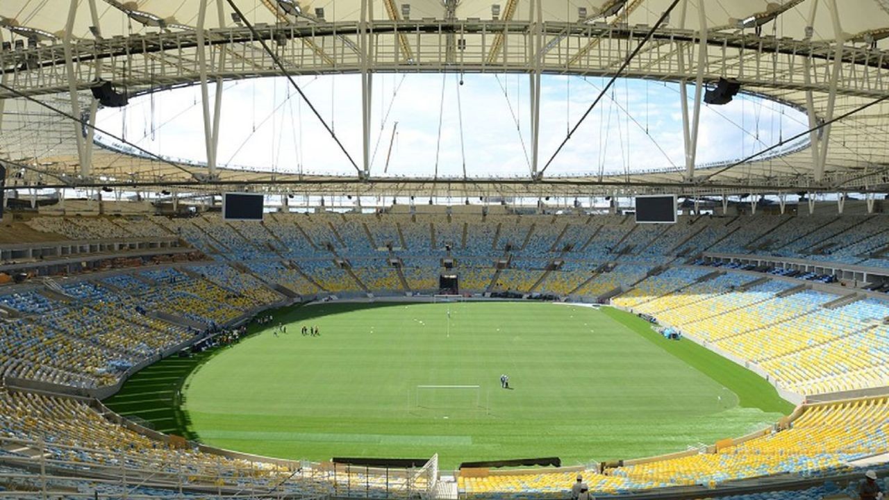 Vista panorâmica do interior do Maracanã com arquibancadas vazias em tons de azul e amarelo, gramado verde e teto retrátil branco com estrutura metálica aparente. Telões escuros estão visíveis sob a cobertura. Pequenos grupos de pessoas estão no campo.