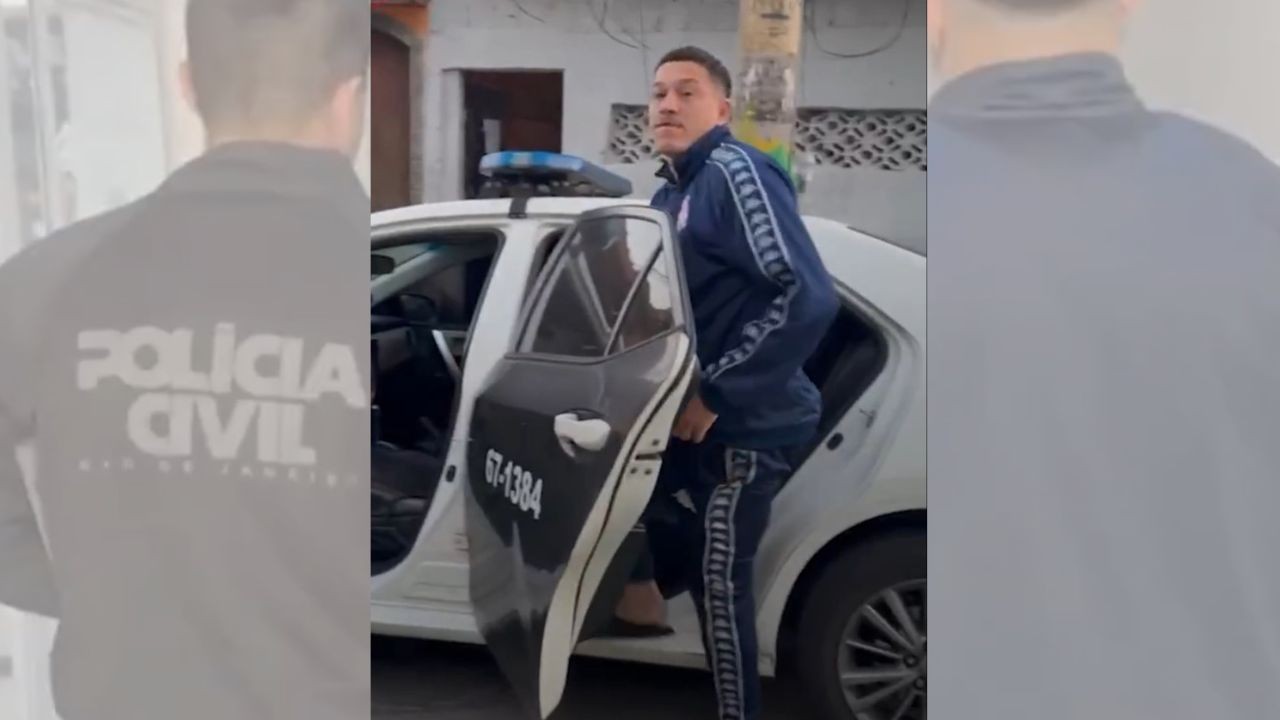 Homem de cabelo curto, vestindo agasalho azul com listras brancas, saindo de um carro da Polícia Civil com a porta aberta. Ao fundo, um policial de costas com POLÍCIA CIVIL estampado na camisa
