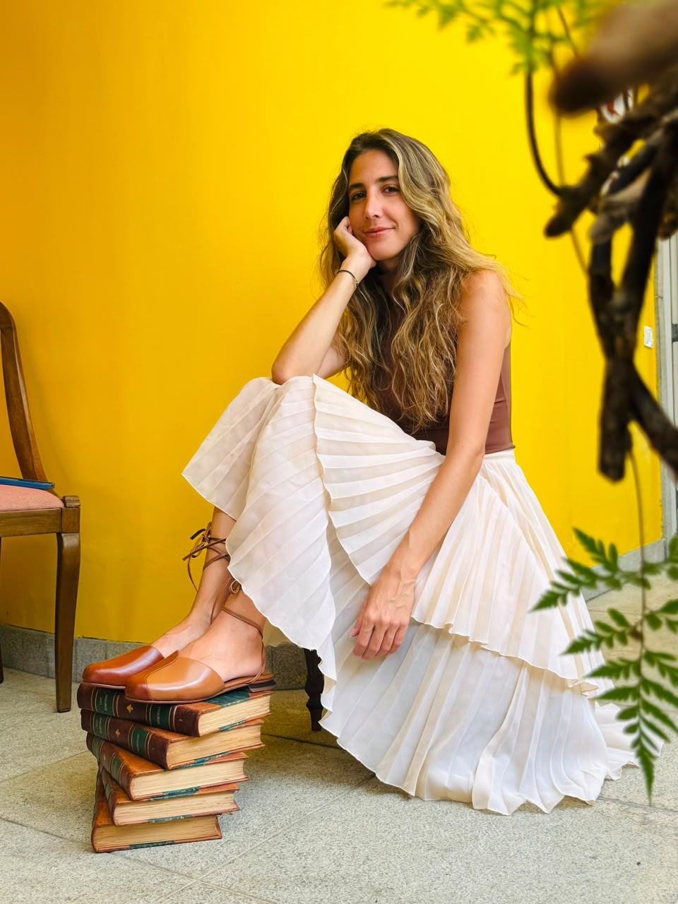 Mulher sorridente com cabelos longos e ondulados, usando blusa marrom e saia plissada bege, sentada no ch&atilde;o com os p&eacute;s sobre uma pilha de livros antigos. O fundo &eacute; uma parede amarela vibrante, com folhagens verdes no canto superior direito e uma cadeira de madeira &agrave; esquerda
